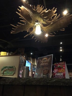 Inside look of Timberwood Grill, it has a huge antlers of elk or moose as a ceiling light and then it has several posters showing some deals at Timberwood Grill